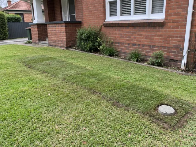 Backfilled lawn trench beside a brick house with a capped sewer access cover in Roseville